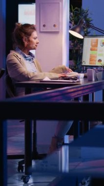Dikey video: İş kadını iş yerinde sabit telefondan konuşuyor, mali yatırımı çözmek için ofis telefonuna cevap veriyor. Girişimci, mesai saatleri dışında şirket işindeki telefon hattını kullanıyor. El bilgisayarı