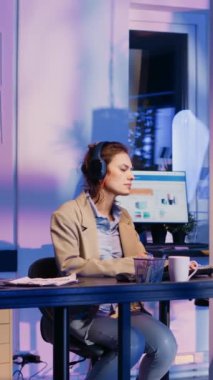 Vertical video: Happy advisor working on business report with headset, listening to music and checking economy diagrams on pc. Female worker planning startup progress before deadline, wearing