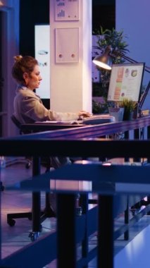 Vertical video: Happy entrepreneur analyzing online statistics late in company office, planning marketing development at night. Young adult looking at economy diagrams to create official report