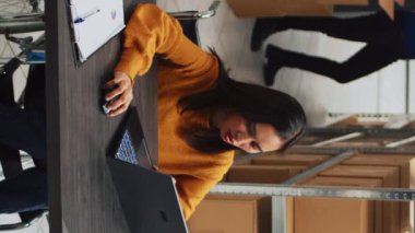 Dikey video: Depoda çalışan ve dizüstü bilgisayarında stok lojistiği ve envanter işleri yapan genç bir çalışan. Kadın tekerlekli sandalye kullanıcısı maluliyet dostu planlama yapıyor