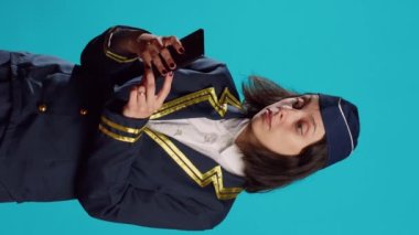 Vertical video: Aircrew member browsing online website on camera, using smartphone for social media app. Young air hostess in flying uniform playing with mobile phone and online browser over blue
