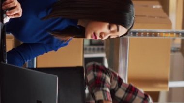 Vertical video: Team of people preparing goods for shipment, checking packages before sending retail store orders. Young man and woman looking at merchandise in packs, storage room shelving. Handheld