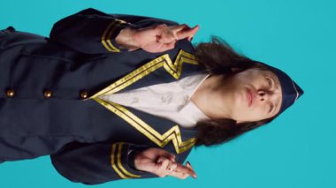 Vertical video: Positive air hostess standing with fingers crossed on camera, asking to fulfill wish over blue backdrop. Female aircrew member praying for luck and fortune, acting hopeful and wishful.