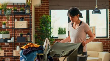 Young adult using steam iron to smooth out wrinkles on clothing, ironing pile of laundered clothes on ironing table. Casual person doing spring cleaning at home, housekeeping work.