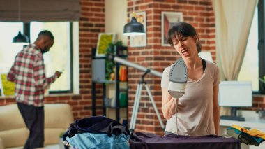 Giysileri ütülerken gülümseyen ve dans eden kişi, ev hanımı evde ev işlerini yaparken eğleniyor. Neşeli eş kıyafetleri ütülemekten ve müzik dinlemekten zevk alıyor. El kamerasıyla..