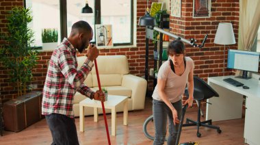 Smiling young people using tools to clean household, listening to music and dancing while they use mop and vacuum. Diverse couple cleaning apartment floors, sweeping dust with solution.