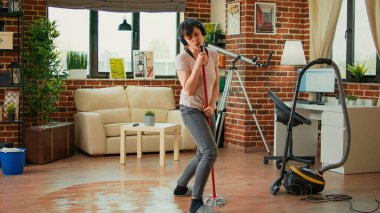 Cheerful girlfriend listening to music on headset and mopping floors, sweep dirt. Housewife feeling happy and cleaning household with tools and appliances, enjoying dance moves.