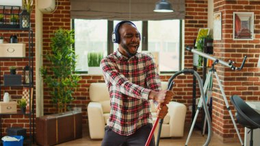 Cheerful funny boyfriend using mop to clean wooden floors, cleaning household and listening to music on headphones. Young smiling man enjoying mopping to clean dirt, household chores.