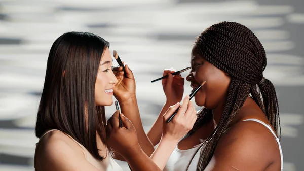 Diverse women playing with make up brushes in studio, giving eachother makeovers and having fun as friends. Young beautiful girls enjoying skincare and body positivity campaign.