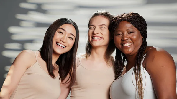 Multiethnic group of models posing for body positivity ad in studio, having fun with friends advertising skincare products. Cheerful women laughing on camera, different skintones and body types.