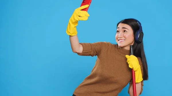 Cheerful housekeeper holding cleaning broom while taking selfie with smartphone, standing in studio over blue background. Smiling maid using high-quality detergents and rubber gloves