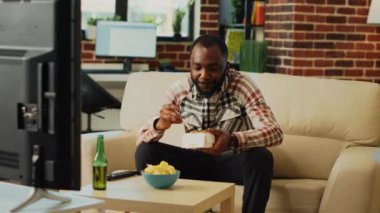 African american man using chopsticks to eat noodles, drinking beer and watching movie on tv channel program. Person feeling relaxed at home eating asian takeaway meal by himself.