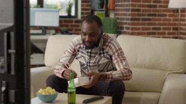 Happy guy eating noodles using chopsticks at home, opening bottle of beer and feeling relaxed at television. Smiling adult enjoying asian delivery meal for dinner, watching show on tv.