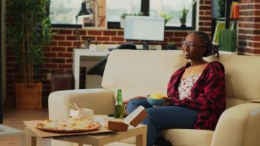 Happy woman laughing at favorite film on tv and drinking beer, holding bowl of chips in living room. Young adult enjoying movie on television with snacks and drinks, takeaway meals.