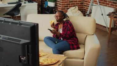 Cheerful woman eating slice of pizza and watching film, having fun with favorite tv show and food delivery meal. Young person enjoying takeout order and bottles of beer, watch movie.