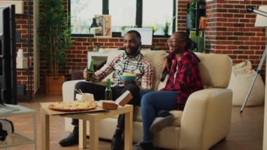 African american couple having fun watching television, ordering burgers, pizza and nodles from delivery takeaway. Young partners enjoying movie or tv show, drinking bottles of beer.