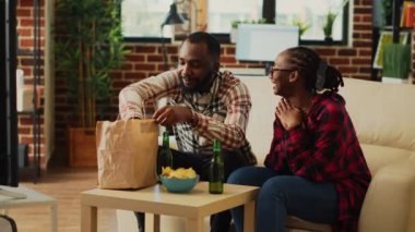 Relaxed people eating hamburgers together at home, enjoying cheeseburgers and fries with bottles of beer. Life partners having fun watching movie on television, drinking alcohol.