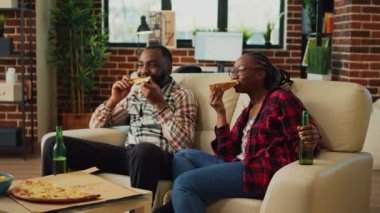 Boyfriend and girlfriend eating pizza from takeaway, having fun with fast food and beer in living room. Partners feeling relaxed in front of television, watching movies at home.