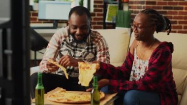 Happy life partners eating slices of pizza on couch, having fun together watching favorite movie on television. Young man and woman in relationship enjoying delivery food and alcohol.