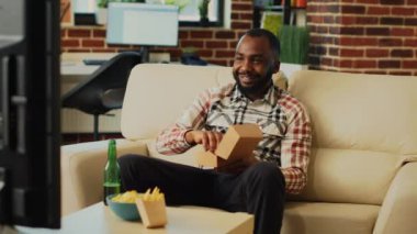 Modern guy eating cheeseburger with fries from takeout, enjoying fast food delivery meal and watching comedy movie on television. Young happy adult feeling relaxed in front of tv.
