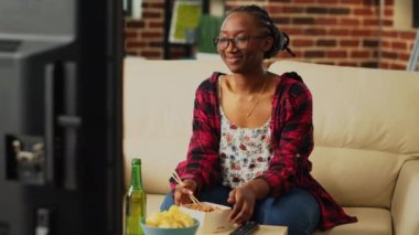 Cheerful girl using chopsticks to eat noodles, laughing at favorite comedy movie on television. Young adult eating asian food in takeaway box and drinking alcohol, having fun at tv.