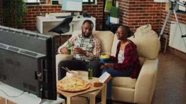 African american people watching film on tv together, drinking beer and eating chips on sofa. Modern couple feeling relaxed with film on television and multiple types of fast food from delivery.