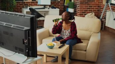 African american girl eating noodles with chopsticks, serving beer and binge watching favorite tv show on sofa. Happy woman enjoying asian meal from takeout delivery order, watch television.