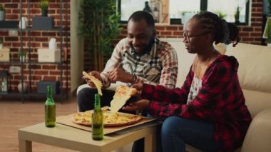 Happy partners eating slices of pizza on sofa and binge watching tv show at home, enjoying fast food meal. Modern couple receiving takeaway food order and sitting to have dinner at television.