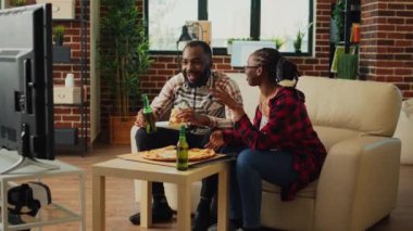 African american couple eating pizza and drinking beer in living room, preparing to watch comedy movie on television. Young people having fun with fast food delivery meal and drinks.