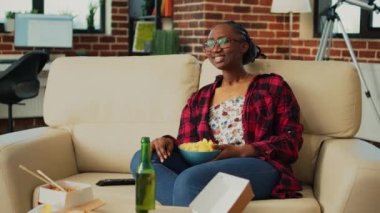 Happy woman laughing at comedy show on television, eating chips and snacks from bowl in living room. Smiling modern girl serving multiple fast food meals from takeout, drinking beer.