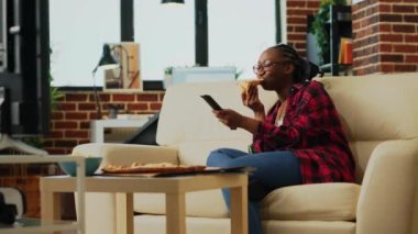 Female adult eating pizza slices from takeaway on sofa, enjoying film on television with alcohol bottles and fast food delivery meal. Cheerful girl having fun watching tv show. Handheld shot.