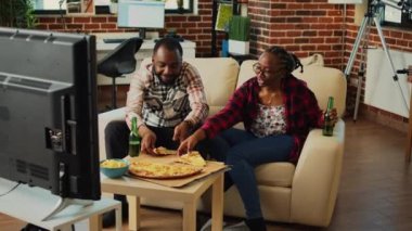 Young cheerful life partners eating slices of pizza in front of television, watching favorite movie or tv show on couch. Relaxed happy people having fast food dinner and drinking beer.