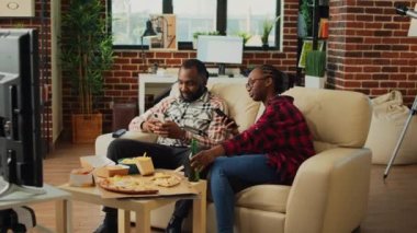 African american people using smartphones on couch, sitting together in front of television and browsing mobile phone apps. Modern partners enjoying leisure time with fast food meal.