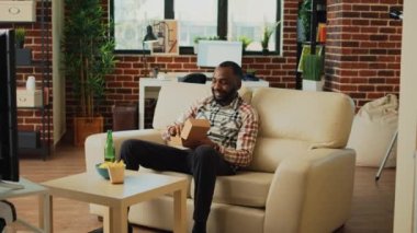 Modern man eating hamburger with fries on couch, having dinner with fast food and beer bottle at home. Young guy feeling relaxed watching comedy or action movie, having fun with tv.