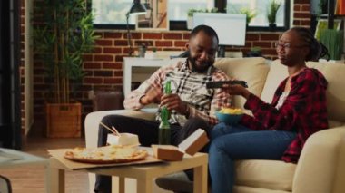Cheerful couple watching favorite show at home, holding bottle of alcohol and eating snacks in bowl. Man and woman in relationship feeling happy with film on television and chips. Tripod shot.