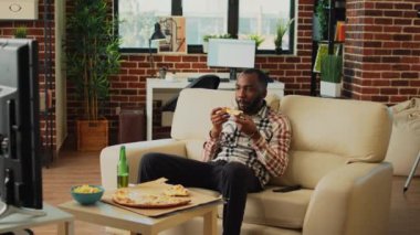 Relaxed guy taking bite of slice of pizza in living room, eating fast food delivery meal and drinking beer from bottle. Young man having fun watching comedy movie on television, eat snacks.