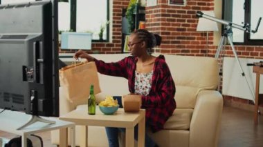 Cheerful person eating hamburger from fast food takeout, enjoying meal with burger and fries while she watches film or comedy show on tv. Woman serving takeaway delivery food at home.