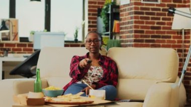 Happy woman switching channels with tv remote control, searching for good movie to watch and eat takeaway meal. Young person browsing television pograms at home, drink beer. Tripod shot.