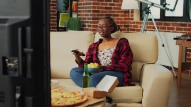 Smiling person holding bowl of snacks watching show, drinking alcohol and eating takeaway meal from multiple fast food orders. Young adult having burgers, fries, pizza and asian food at home.