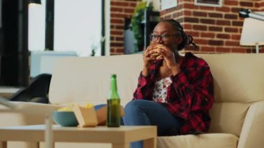 Young woman eating cheeseburger with fries and beer, binge watching favorite tv show by herself at home. Modern girl feeling happy with fast food takeaway, watch film on television. Handheld shot.