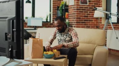 Modern adult having hamburger and fries for dinner, eating fast food meal from takeaway and watching movie on television. Young guy enjoying burger and drinking bottle of beer at home.