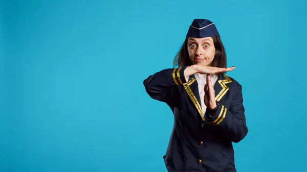 Professional stewardess showing t shape sign in studio, asking for pause and refusing to work. Young confident air hostess in uniform doing break and rejection gesture over blue background.