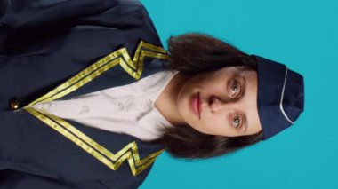 Vertical video: Confident young stewardess posing in flying uniform, feeling happy working in aviation industry on camera. Smiling woman dressed as air hostess looking successful over blue backdrop.