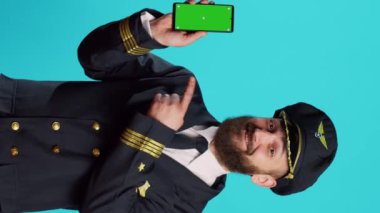 Vertical video: Positive aircrew captain showing phone with greenscreen, holding smartphone with isolated display and chroma key template. Young airplane pilot in uniform looking at mockup copyspace