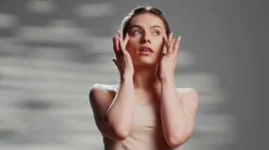 Beautiful young adult applying face cream on camera, creating skincare ad to promote sunscreen and glowing products. Luminous female person using moisturizing serum in studio.