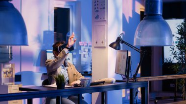 Young worker wearing virtual reality goggles in office, planning financial report to help increase profit. Business woman working late on economy graphs using vr headset at desk.