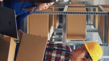 Vertical video: Young man organizing products boxes on racks, doing quality control with business owner in storehouse. Male employee placing packages on shelves, preparing for order distribution.