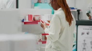 Genç bir kadın eczanede ilaç kutusunu işaretliyor, eczanede ilaçlara bakıyor. Müşteri reçeteli ilaç veya tedavi satın alıyor, uyuşturucu paketleri üzerine broşür okuyor.