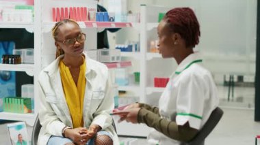 African american specialist showing pack of medicine to young customer, recommending pharmaceutical products in drugstore. Women talking about medicaments and vitamins in pharmacy.