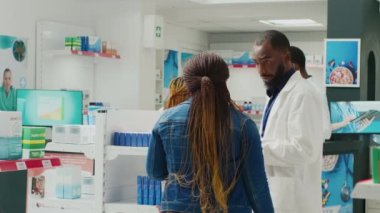 Afro-Amerikalı bir kadın eczacıyla eczane hapları hakkında konuşuyor, ilaç ve uyuşturucuya bakıyor. Kadın müşteri takviye ve vitamin kontrolü yapıyor, eczane danışmanı. Üçayak atışı.